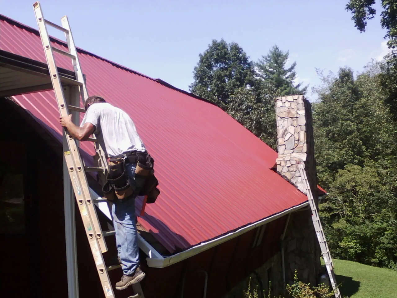 Roofer on a ladder beside a red metal roof and stone chimney — Soffit & Fascia Repair by Roofing Brothers in Maplewood